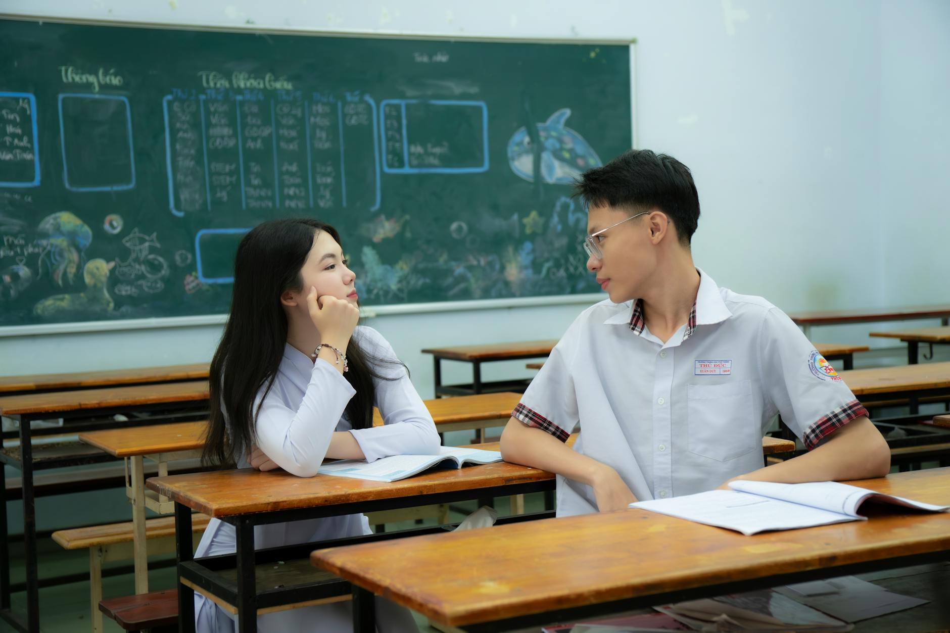 Two students interacting in a classroom setting with books and chalkboard, fostering an academic environment.